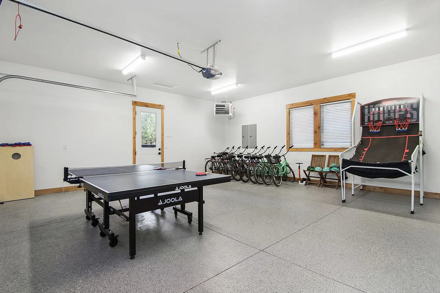 A brightly lit garage game room featuring a ping pong table, an arcade basketball game, and a row of bicycles parked along the wall.