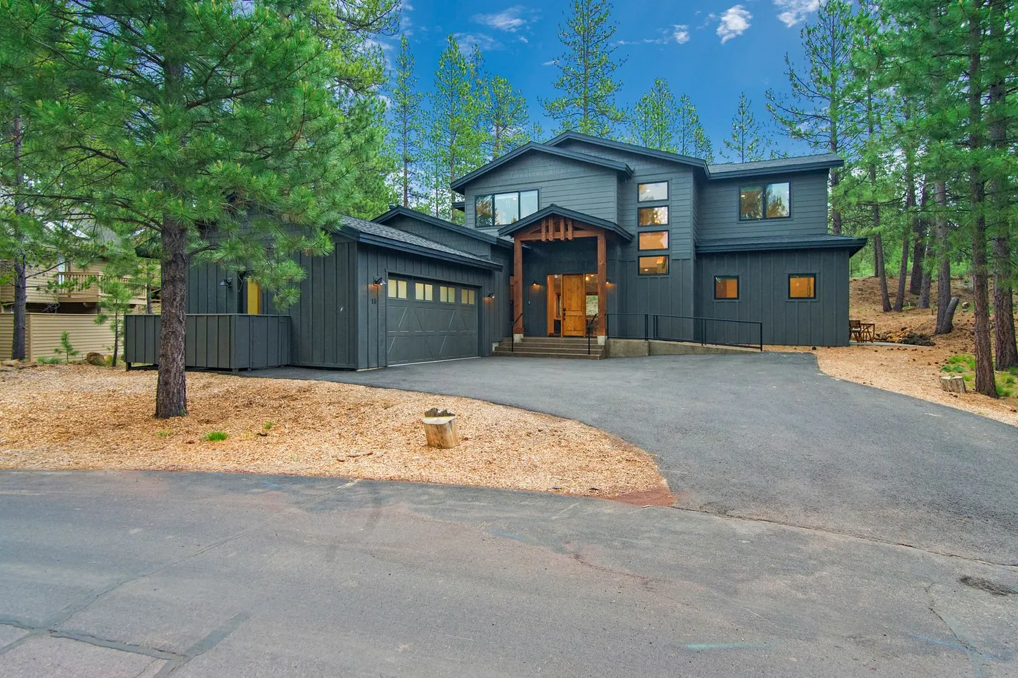 Exterior view of a modern two-story dark gray house with a paved driveway surrounded by pine trees.