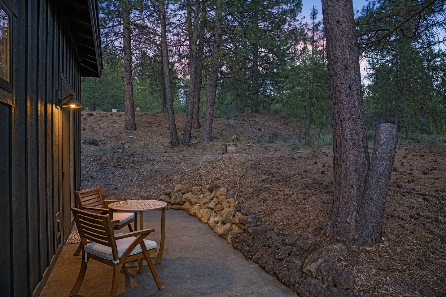 A concrete patio with two wooden chairs and a small table set against a dark siding wall, looking out at a pine forest at dusk.