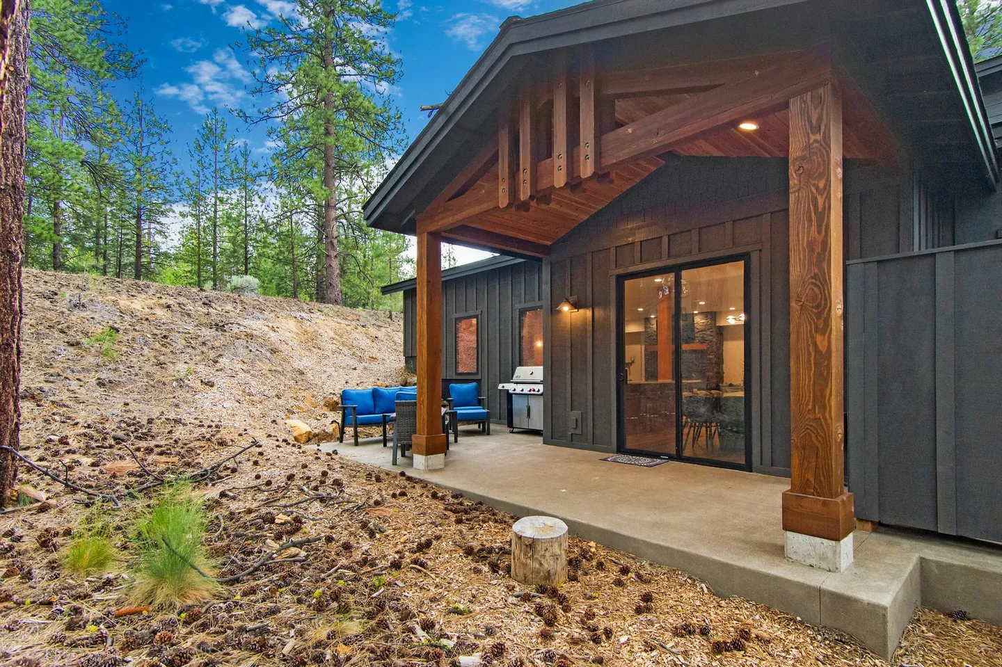 Covered concrete patio with outdoor seating, a barbecue grill, and sliding glass doors leading into the home, surrounded by pine trees.