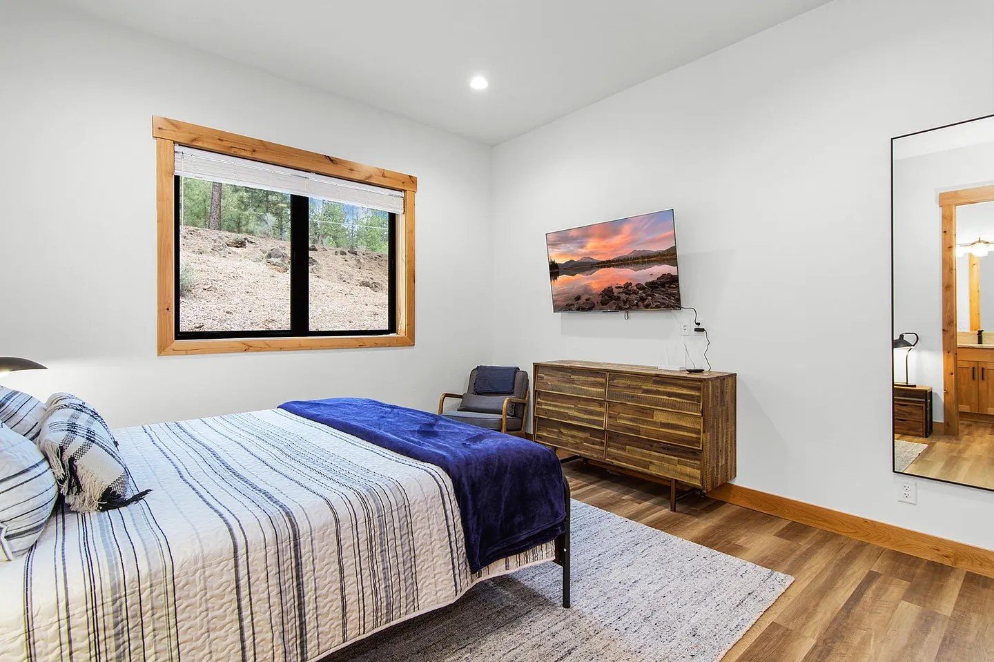 Bedroom with a striped bedspread, wooden dresser, wall-mounted television, and a window with a natural wood frame.