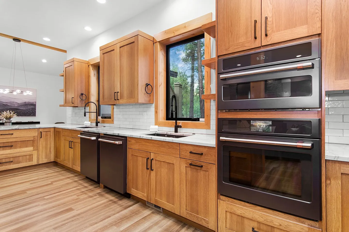 Modern kitchen with natural wood cabinets, dark appliances, built-in double ovens, and large windows looking out to trees.