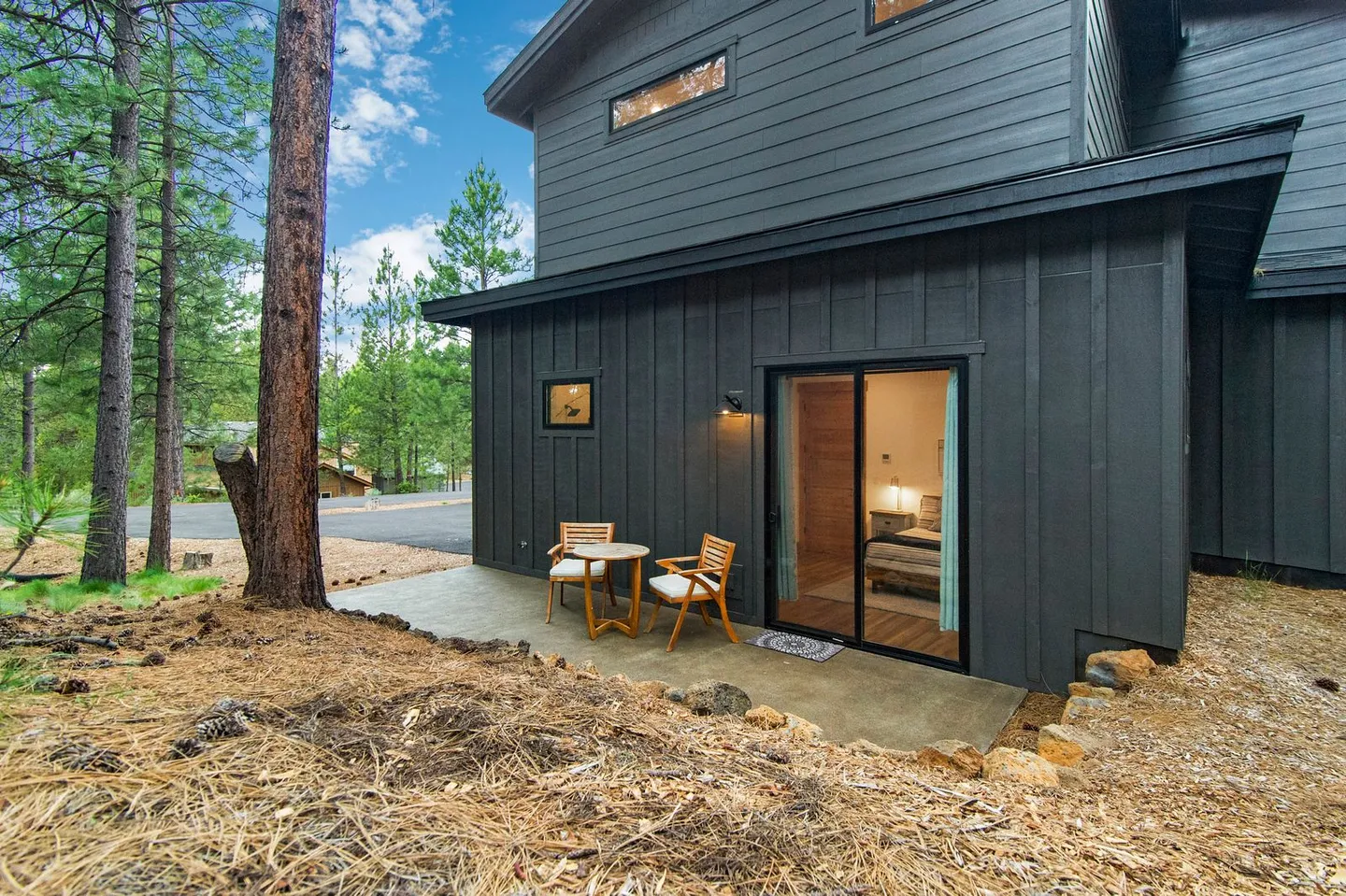 Exterior ground-level concrete patio with a small table and two chairs outside a bedroom sliding glass door of a dark-sided house.