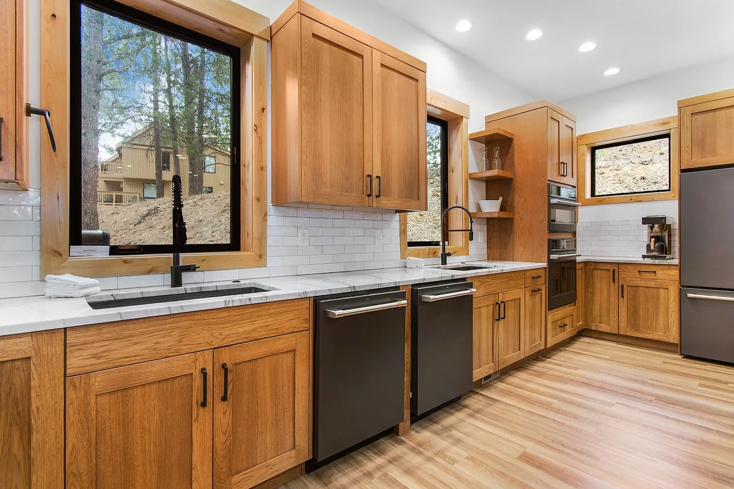 Modern kitchen with natural wood cabinets, light-colored countertops, subway tile backsplash, and two under-mount sinks with black faucets.