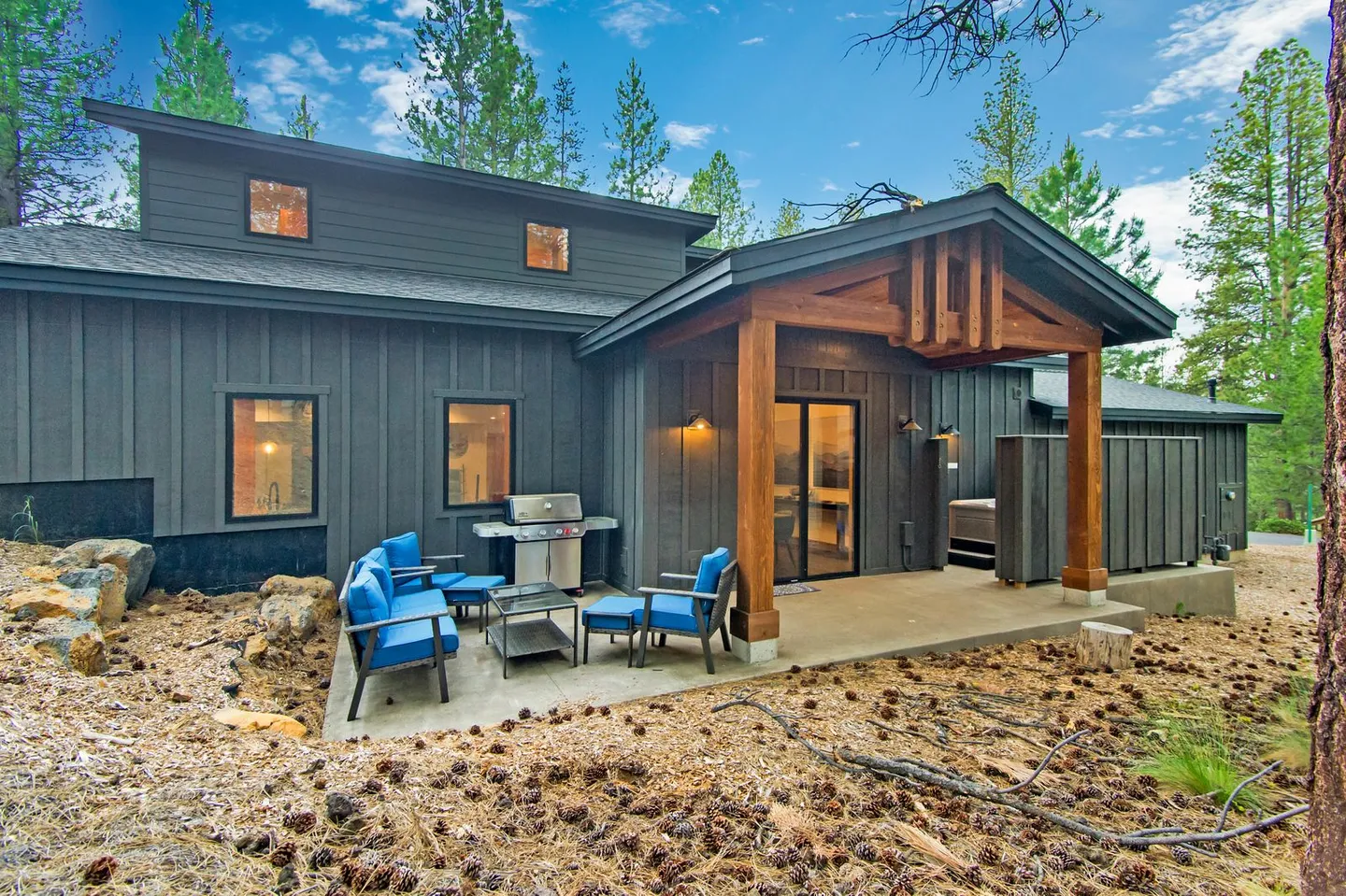 Exterior rear patio with outdoor seating, a barbecue grill, and a covered porch section leading into a dark-sided modern home surrounded by pine trees.