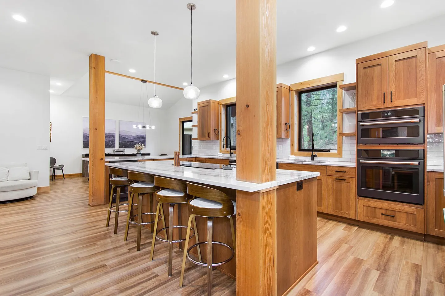 Open-concept modern kitchen with a marble-topped island, barstools, natural wood cabinetry, and double wall ovens.