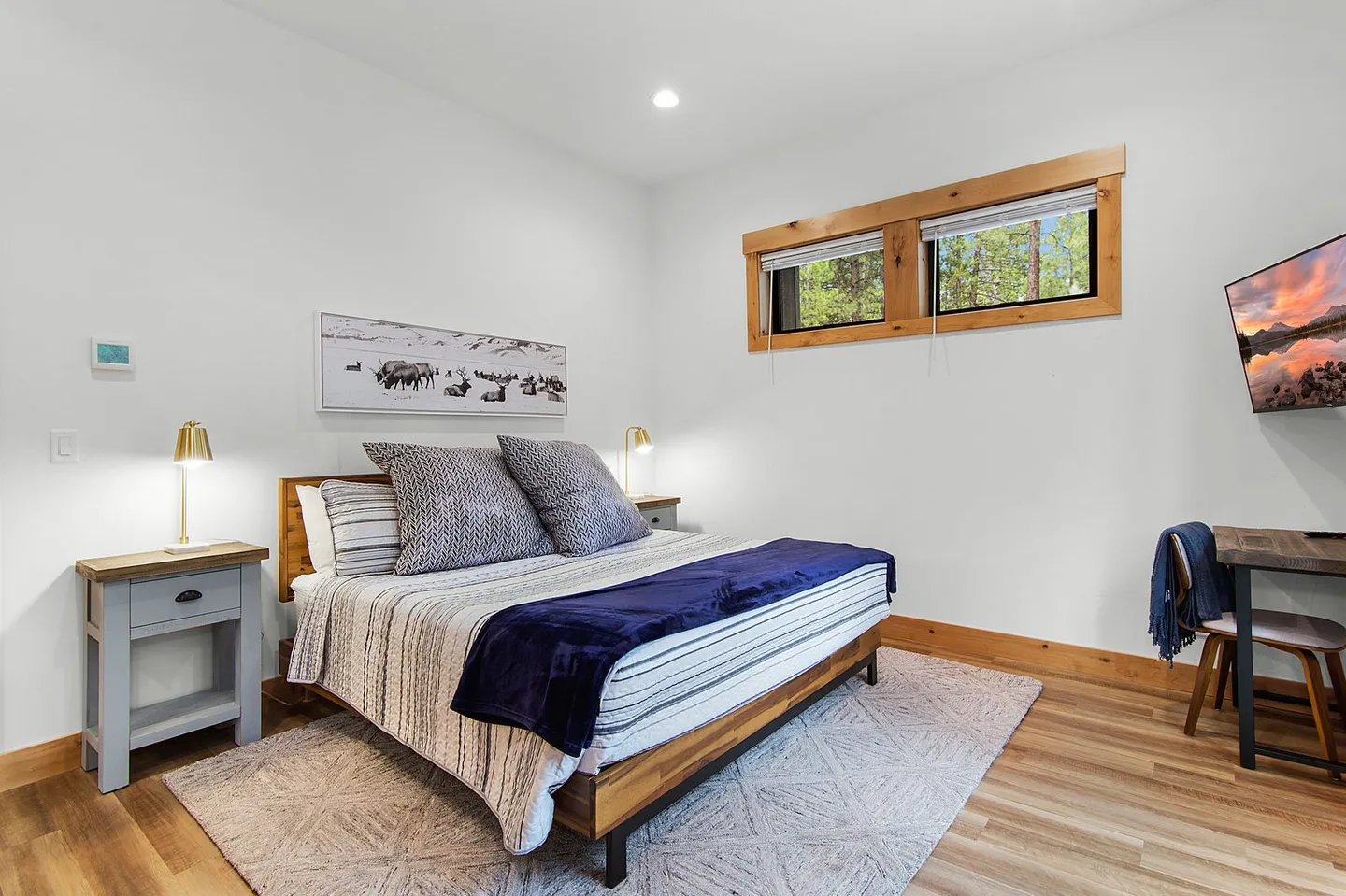 Bedroom with a wooden platform bed, patterned bedding, nightstands with lamps, a wall-mounted TV, and a small desk with a chair.