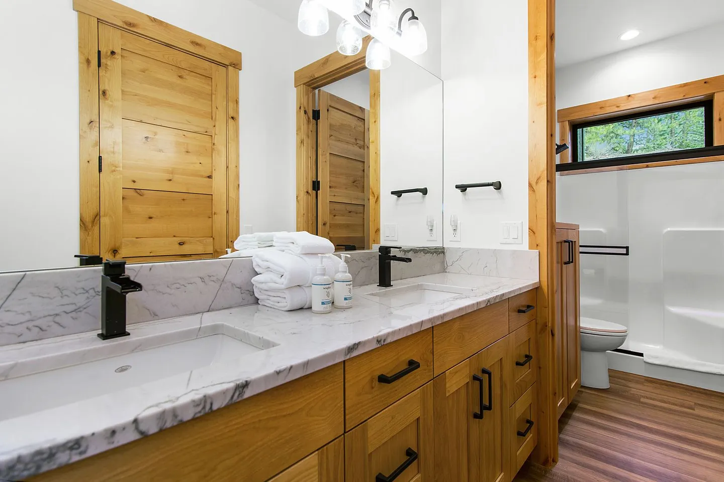 Bathroom featuring a double vanity with light-colored marble-look countertops, wooden cabinets, black fixtures, and a glimpse of the toilet and shower.