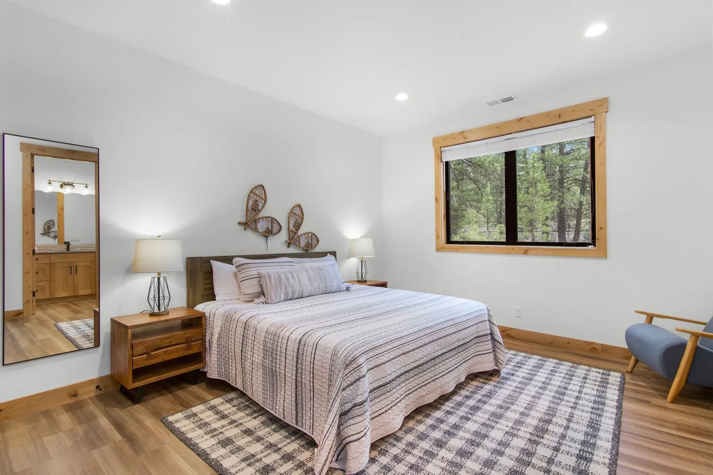 Bedroom featuring a made bed with striped bedding, wood nightstands, a full-length mirror reflecting a bathroom, and a window with wood trim showing trees.