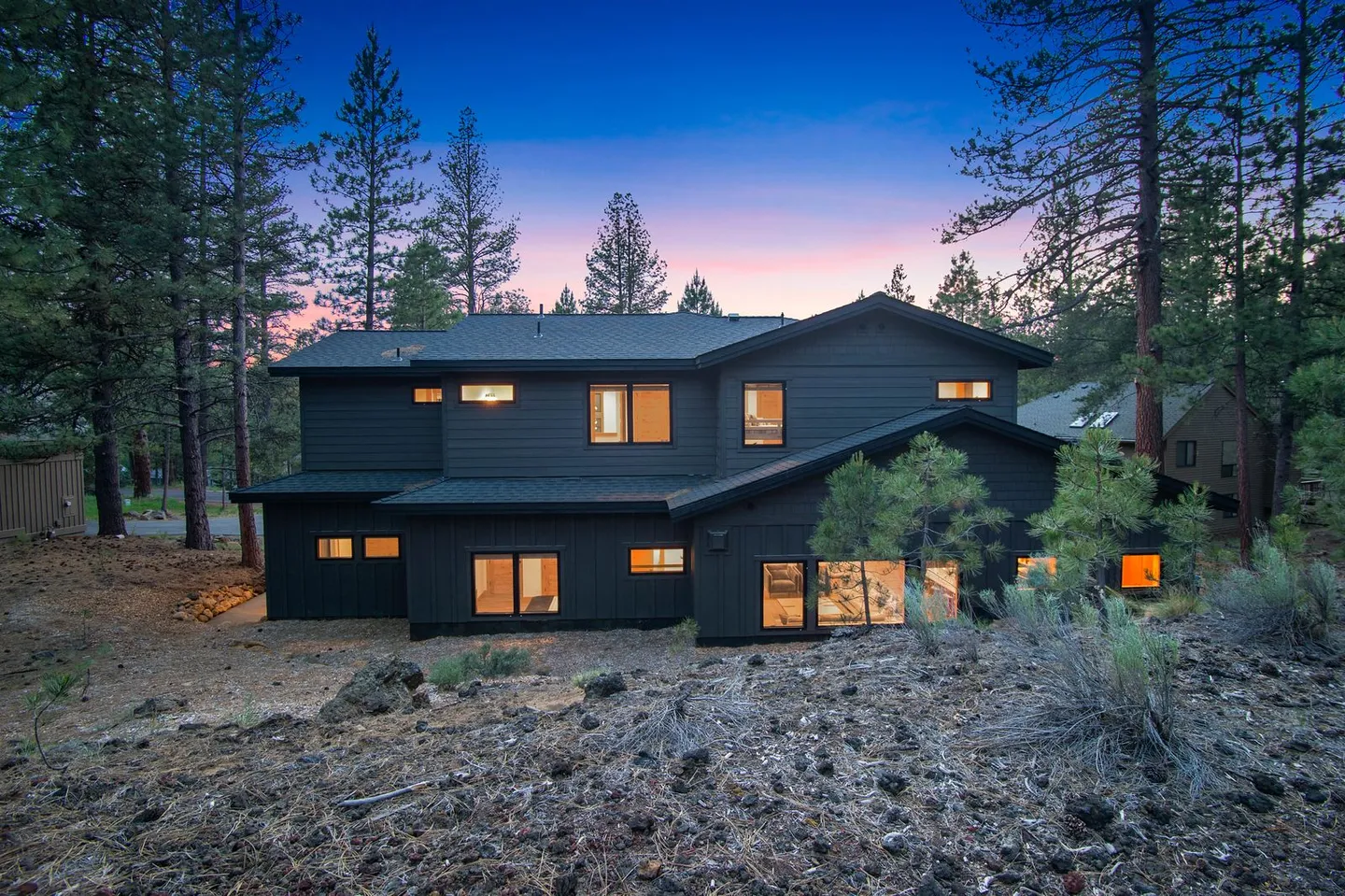 Exterior of a modern dark-sided two-story home surrounded by pine trees at dusk with interior lights glowing.