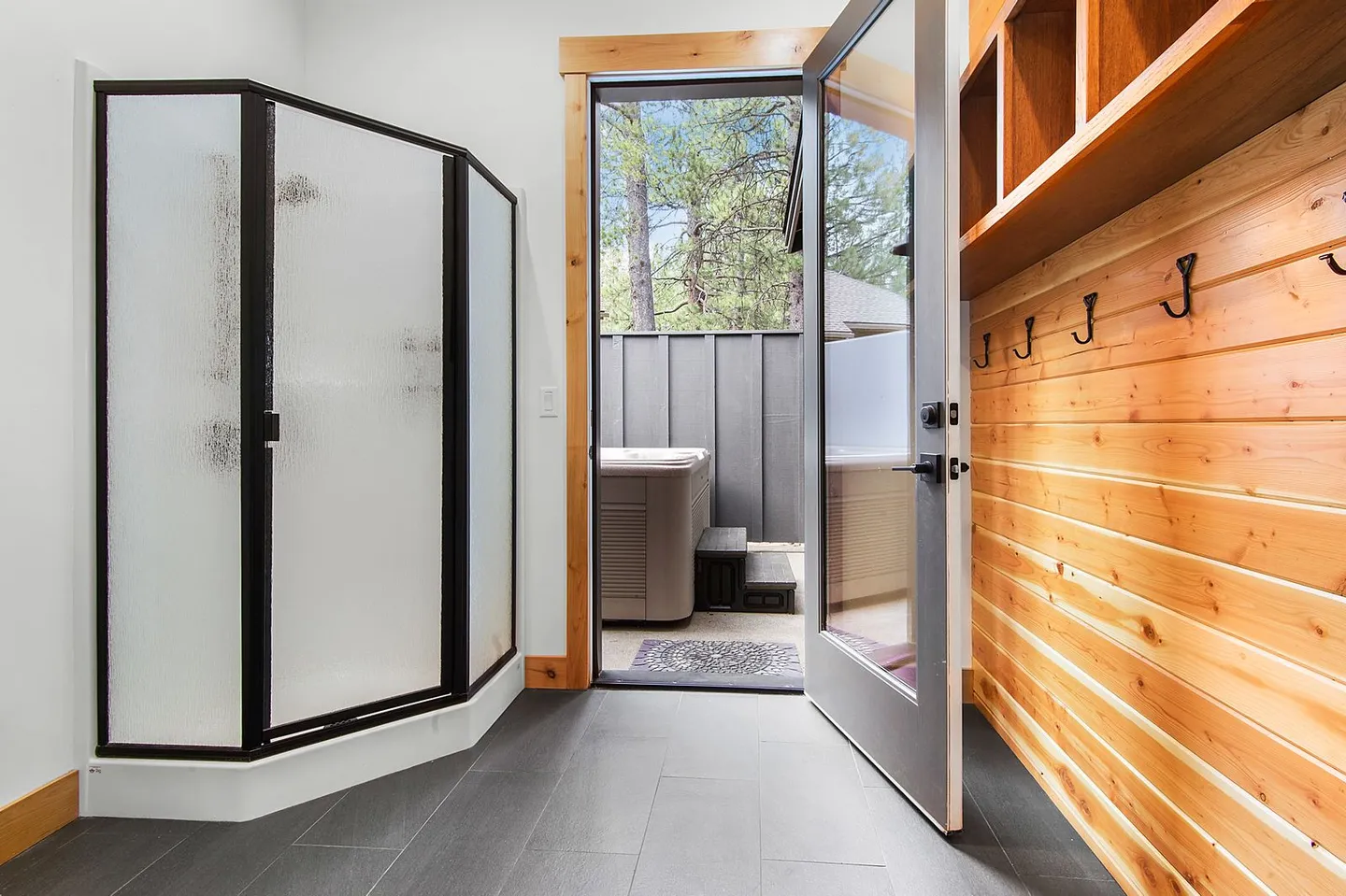 Bathroom with dark tile floor, frosted glass shower stall, wood-paneled wall with coat hooks, and an open glass door leading to an outdoor hot tub area.