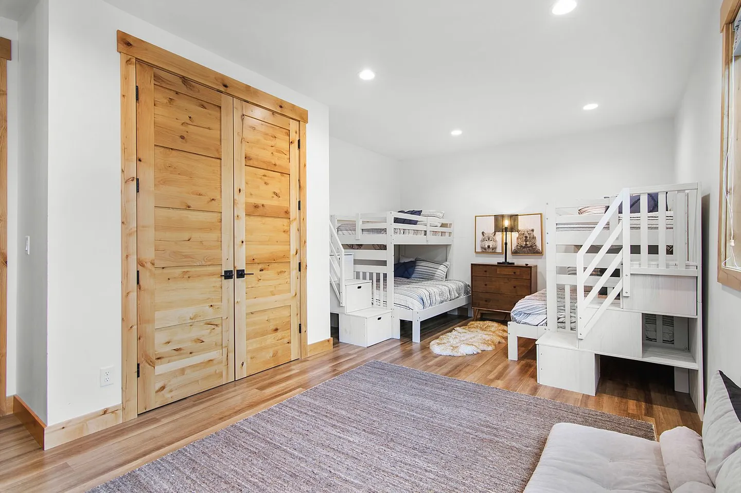 Bunk room featuring two sets of white bunk beds with built-in storage stairs, wood flooring, double knotty wood doors, and recessed ceiling lights.
