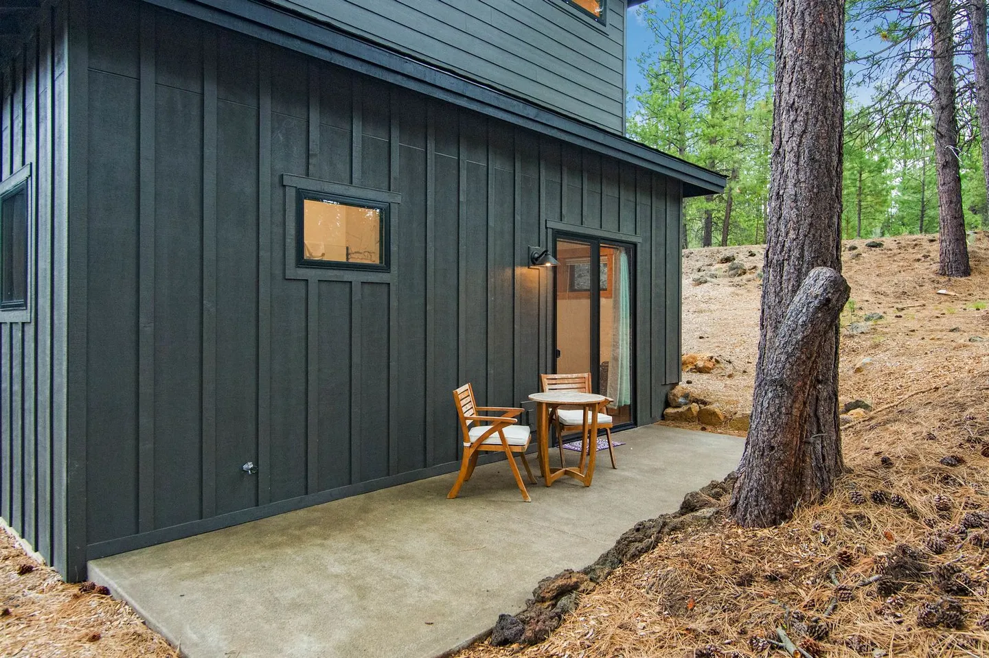 Concrete patio with a wooden table and two chairs outside a dark-paneled house surrounded by pine trees.