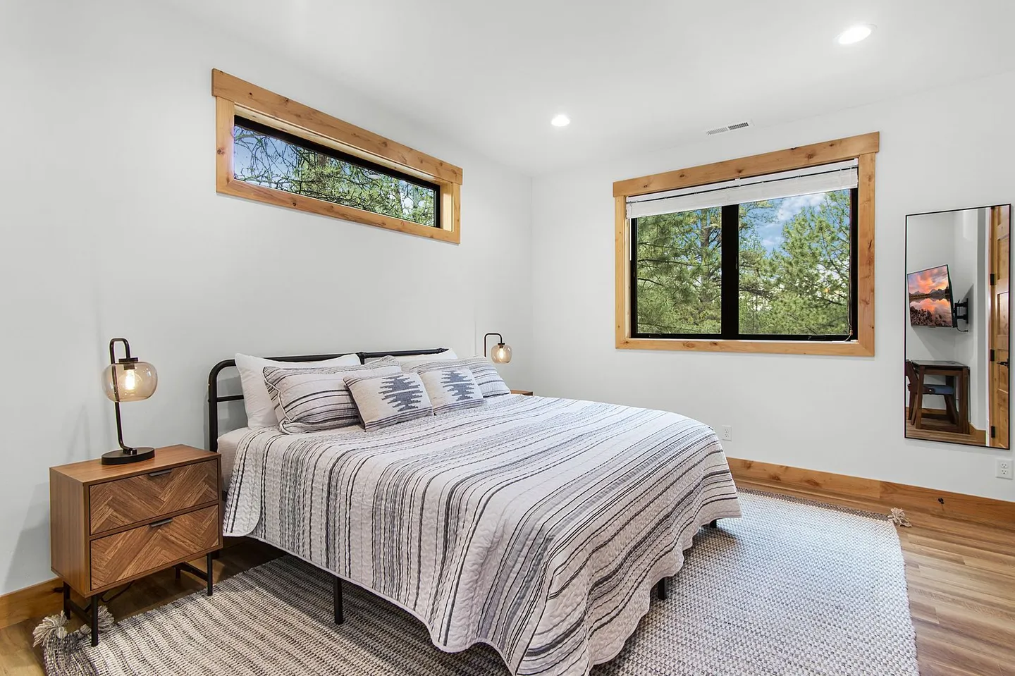 Bedroom with a metal frame bed, striped bedding, wood-trimmed windows, a nightstand, and a full-length mirror.