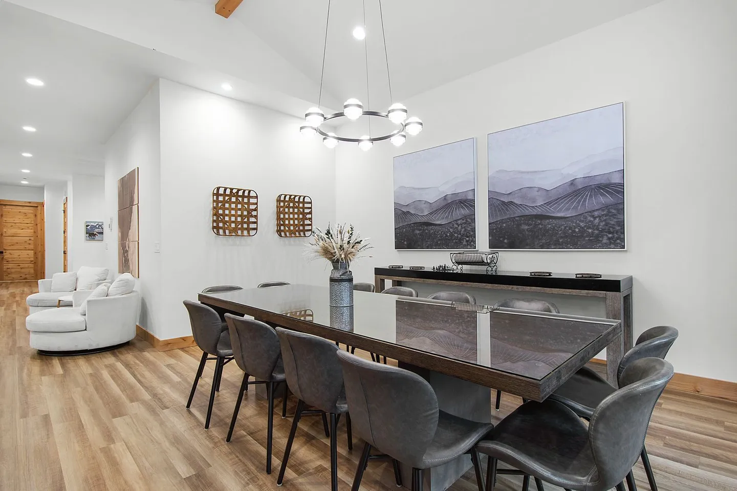 Dining area with a large dining table, gray upholstered chairs, modern ring chandelier, and landscape wall art.