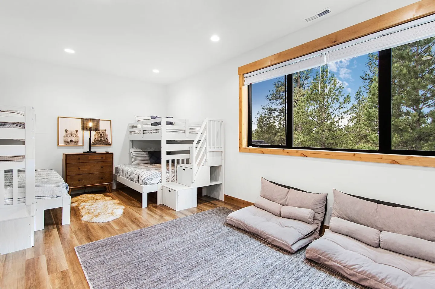 Bedroom with two sets of white bunk beds, a large window overlooking evergreen trees, and low folding lounge cushions on a gray area rug.
