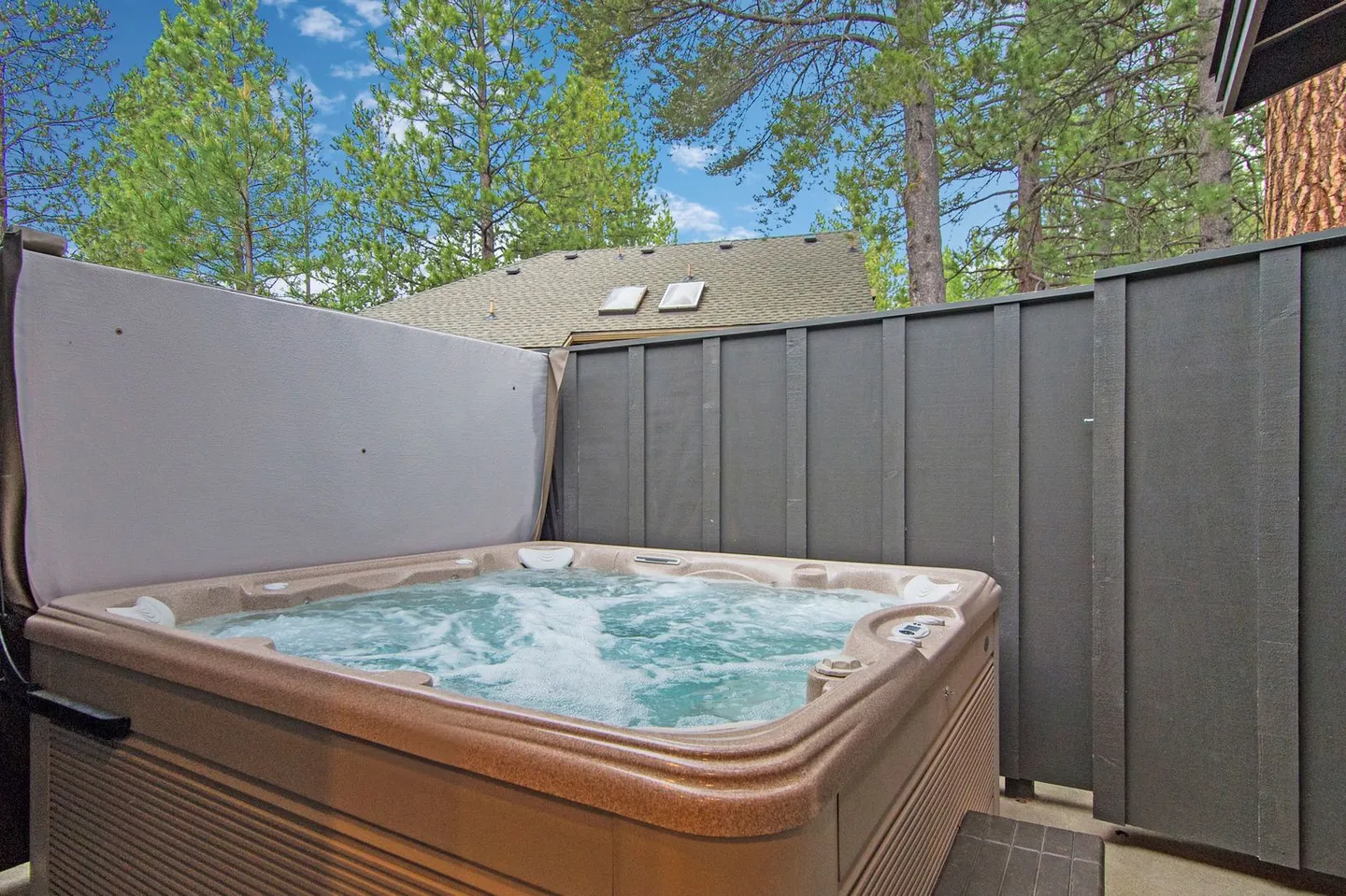 Outdoor bubbling hot tub next to a dark grey privacy fence with pine trees and blue sky above.