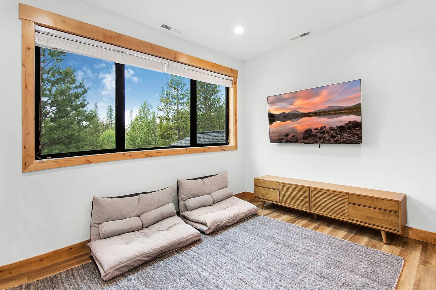 Lounge area with two floor cushions on an area rug, a low wooden media console, a wall-mounted TV, and a large wood-trimmed window showing pine trees.