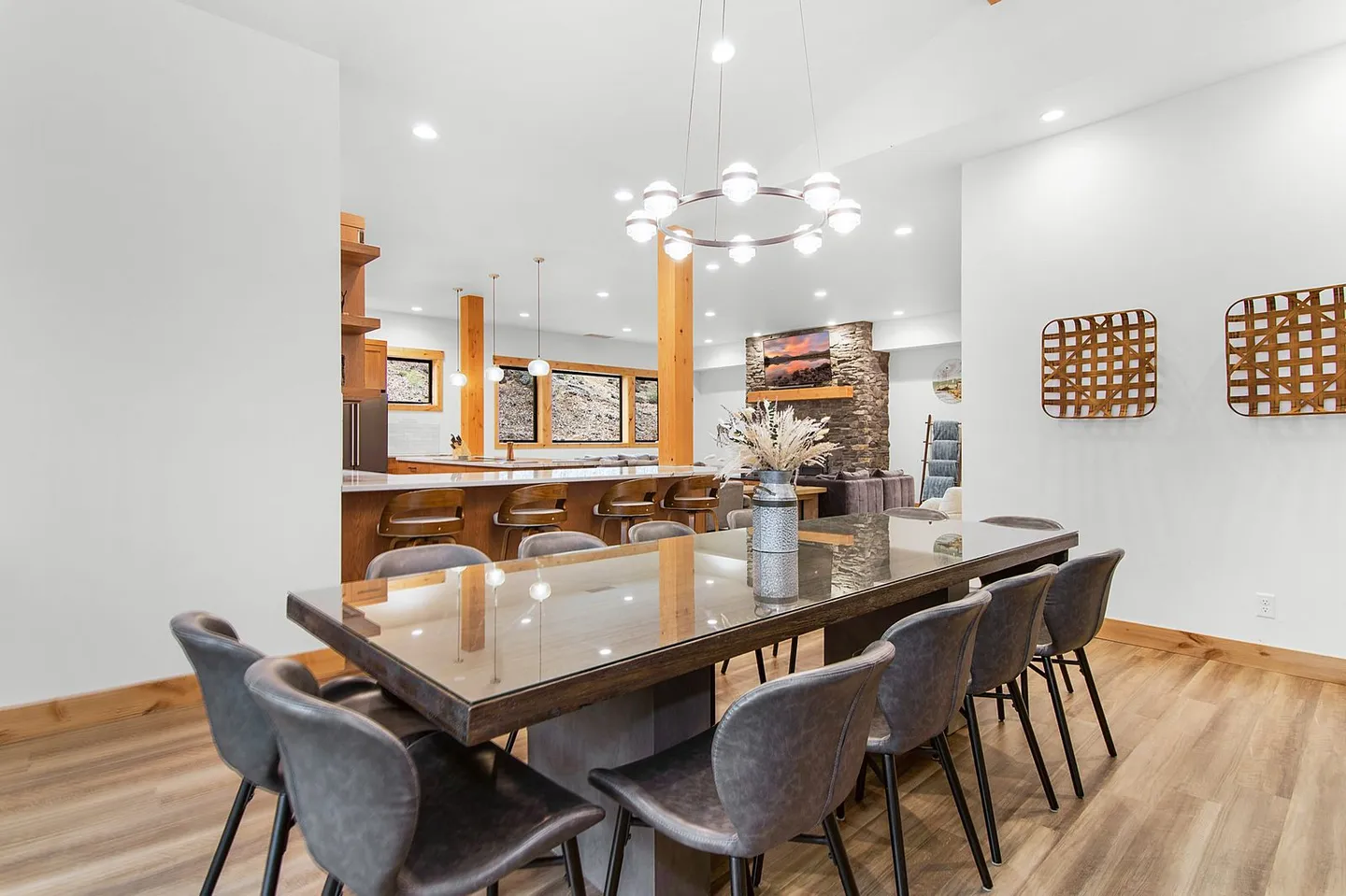 Dining area with a large rectangular table, grey upholstered chairs, and a modern circular chandelier, opening to a kitchen and living space.