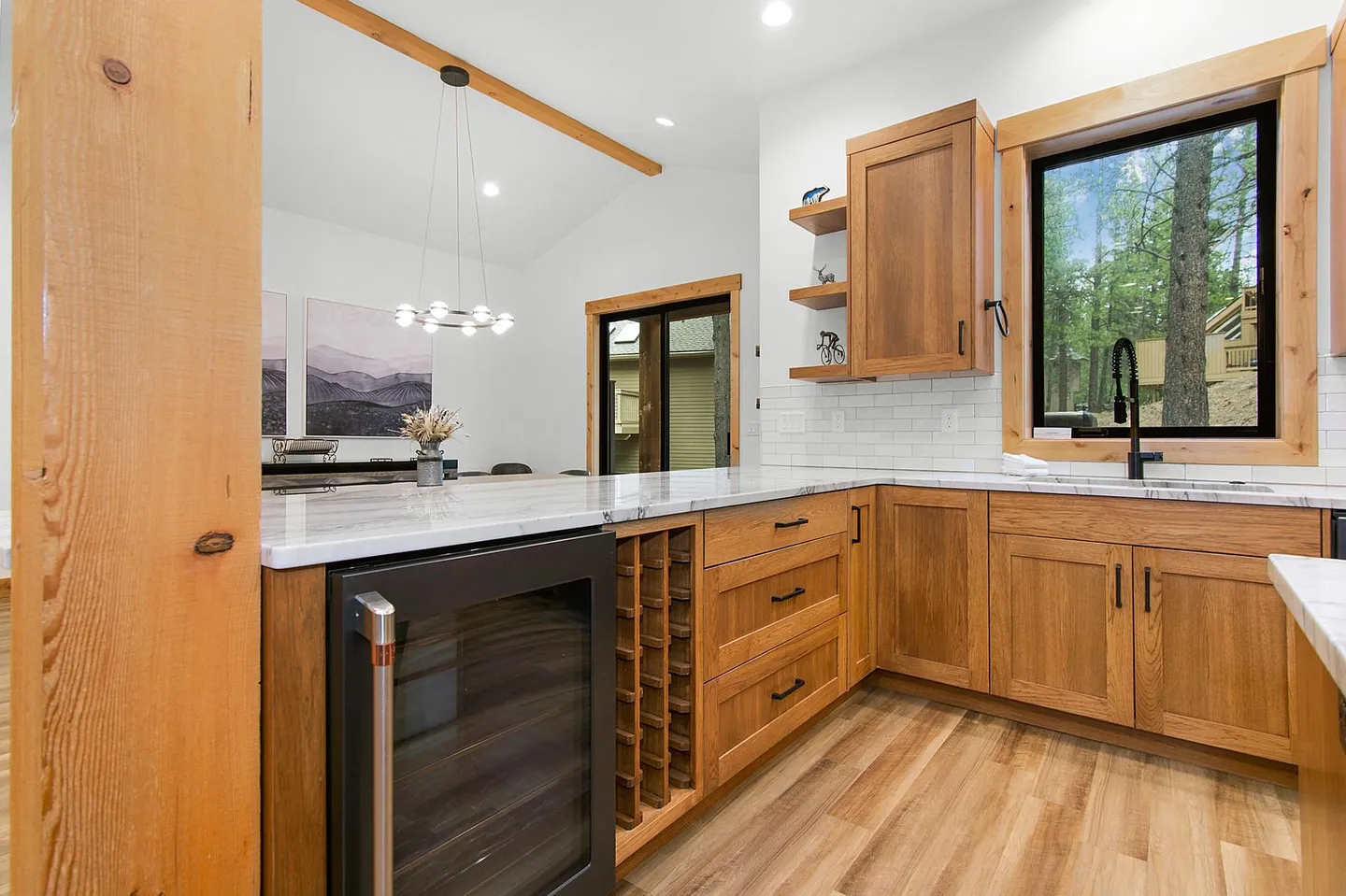 Kitchen counter with light marble surfaces, wooden cabinetry, built-in wine cooler, wine rack, and a window overlooking outdoor greenery.