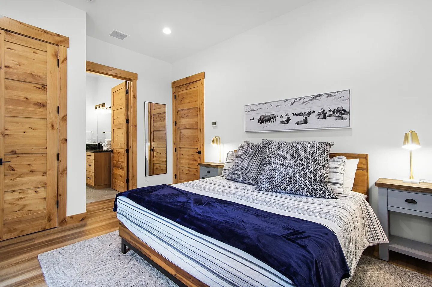 Bedroom with wooden doors, a bed dressed in striped bedding and a blue throw, flanked by grey nightstands with gold lamps, and an ensuite bathroom visible.
