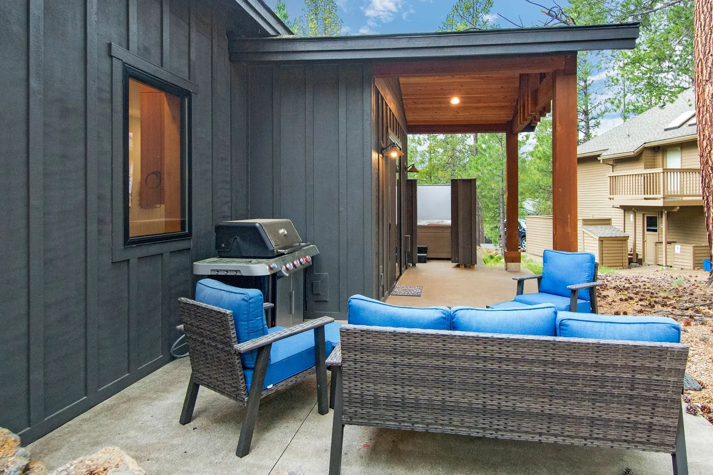 Outdoor concrete patio featuring wicker seating with blue cushions, a barbecue grill, and a covered porch area with a hot tub.