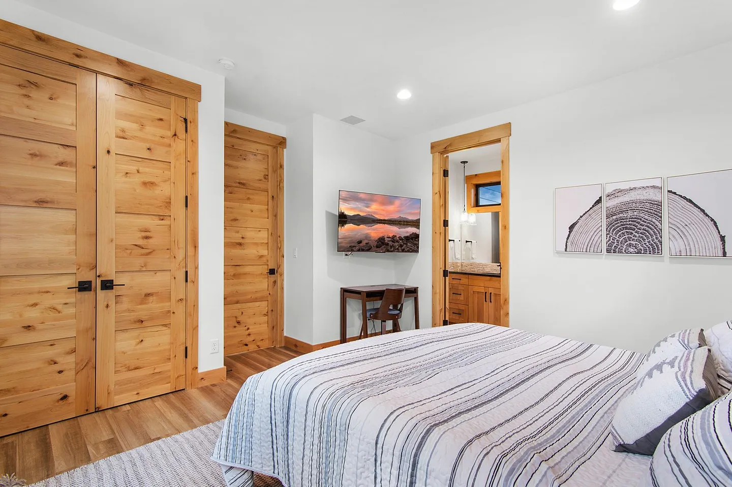 Bedroom with a striped bedspread, light wood doors, a mounted TV, and an open doorway showing an en suite bathroom.