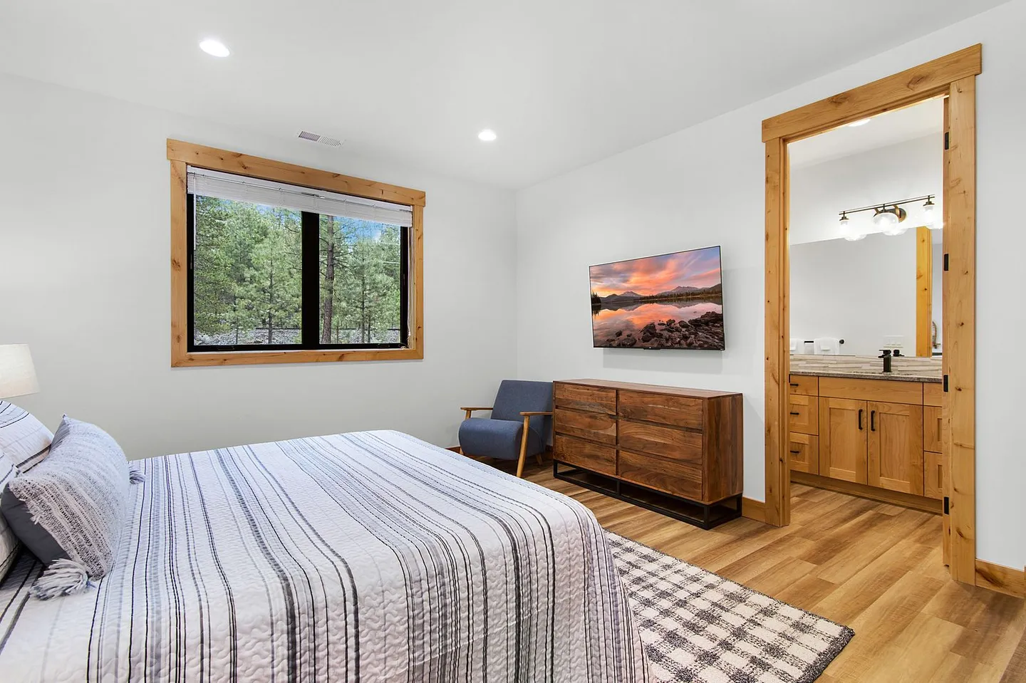 Bedroom with a patterned bedspread, wood-trimmed window overlooking trees, a wall-mounted TV over a wooden dresser, and an open doorway to an ensuite bathroom.