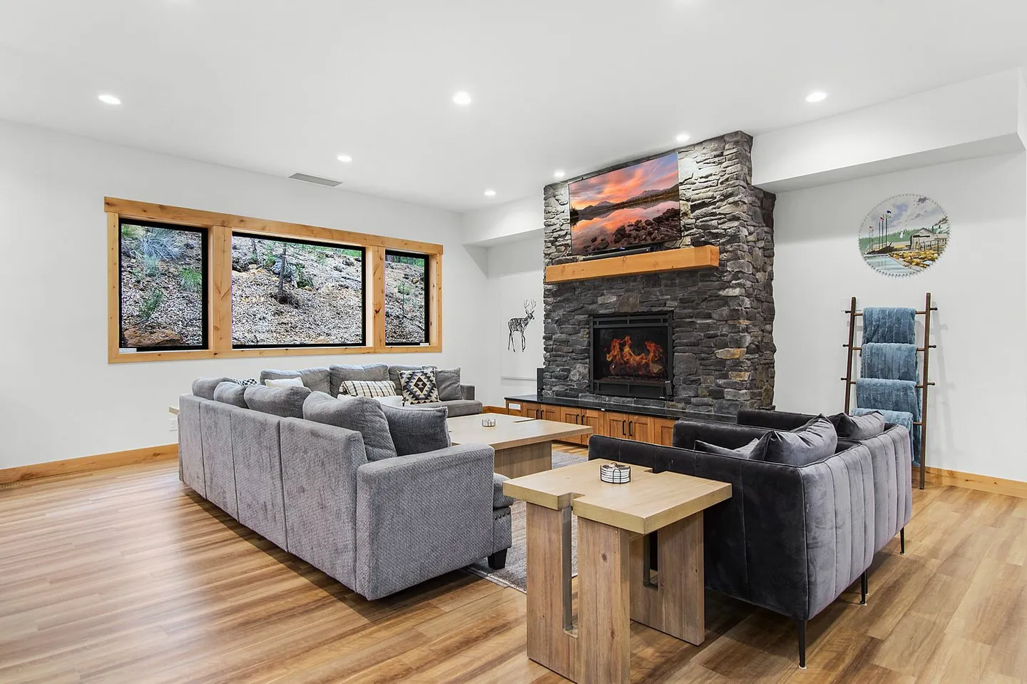 Living room with wood-look flooring, plush sectional sofas, a stone fireplace with a mounted TV, and wood-trimmed windows.