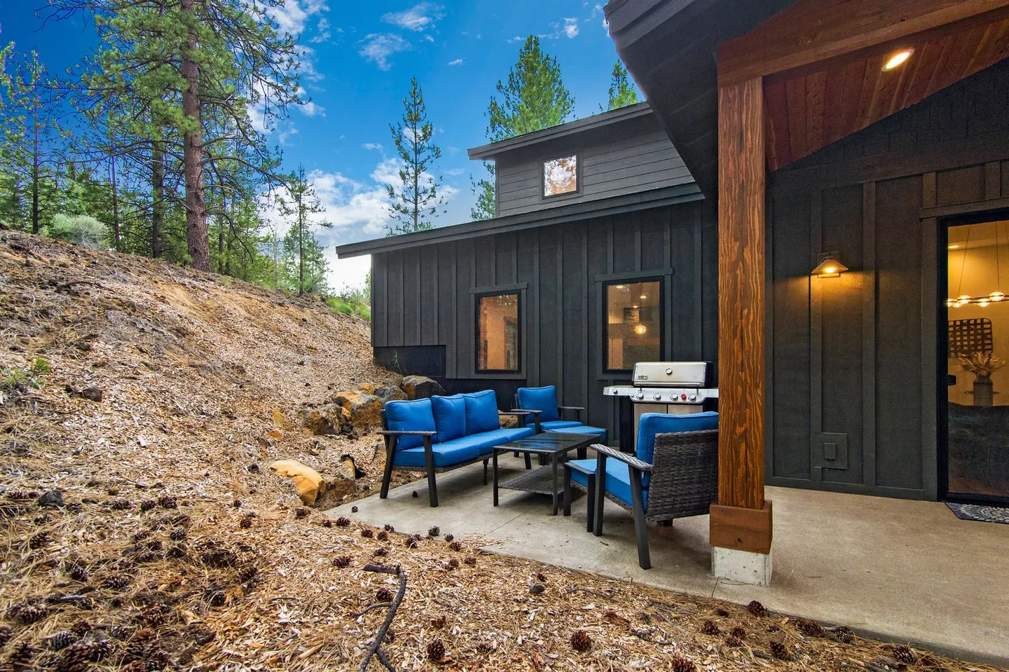 Concrete patio beside a dark-sided house with outdoor blue seating and a barbecue grill next to a sloped hill.