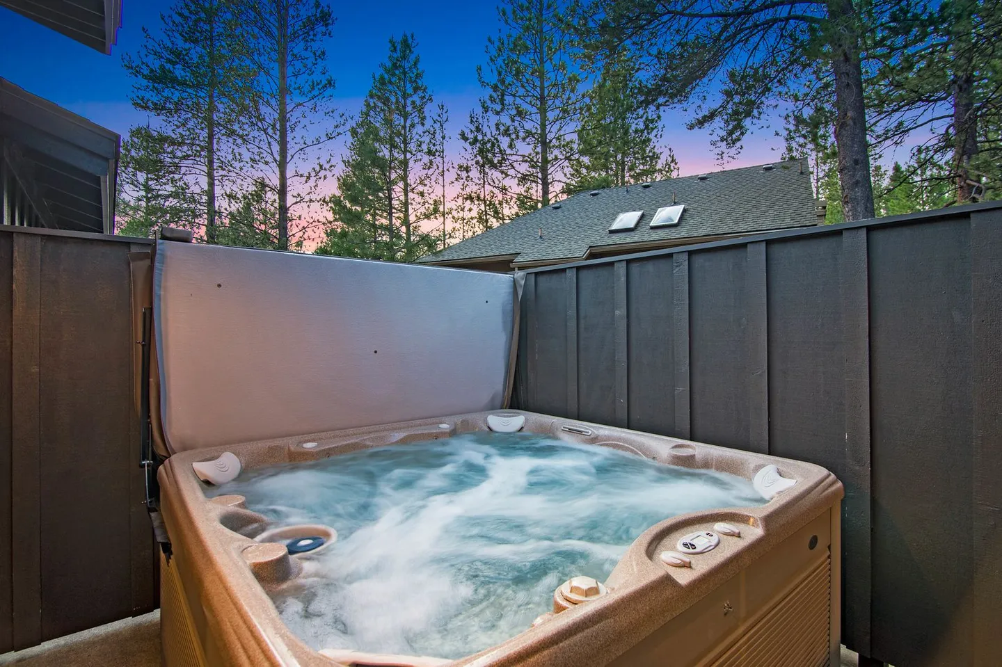 Outdoor hot tub with bubbling water enclosed by dark privacy fences against a backdrop of pine trees at twilight.