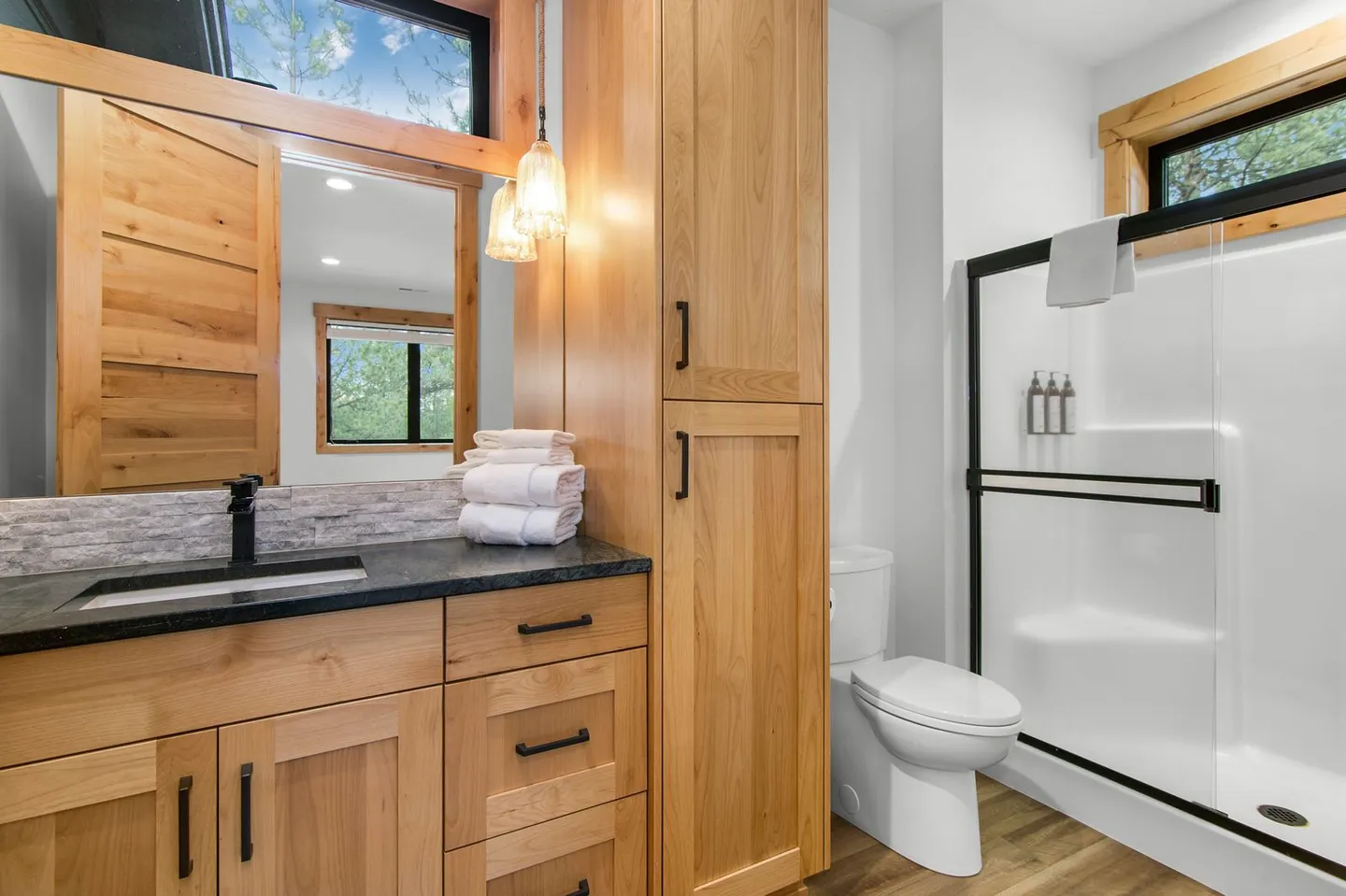 Bathroom featuring a light wood vanity with a dark countertop, matching tall storage cabinet, toilet, and glass-enclosed shower.