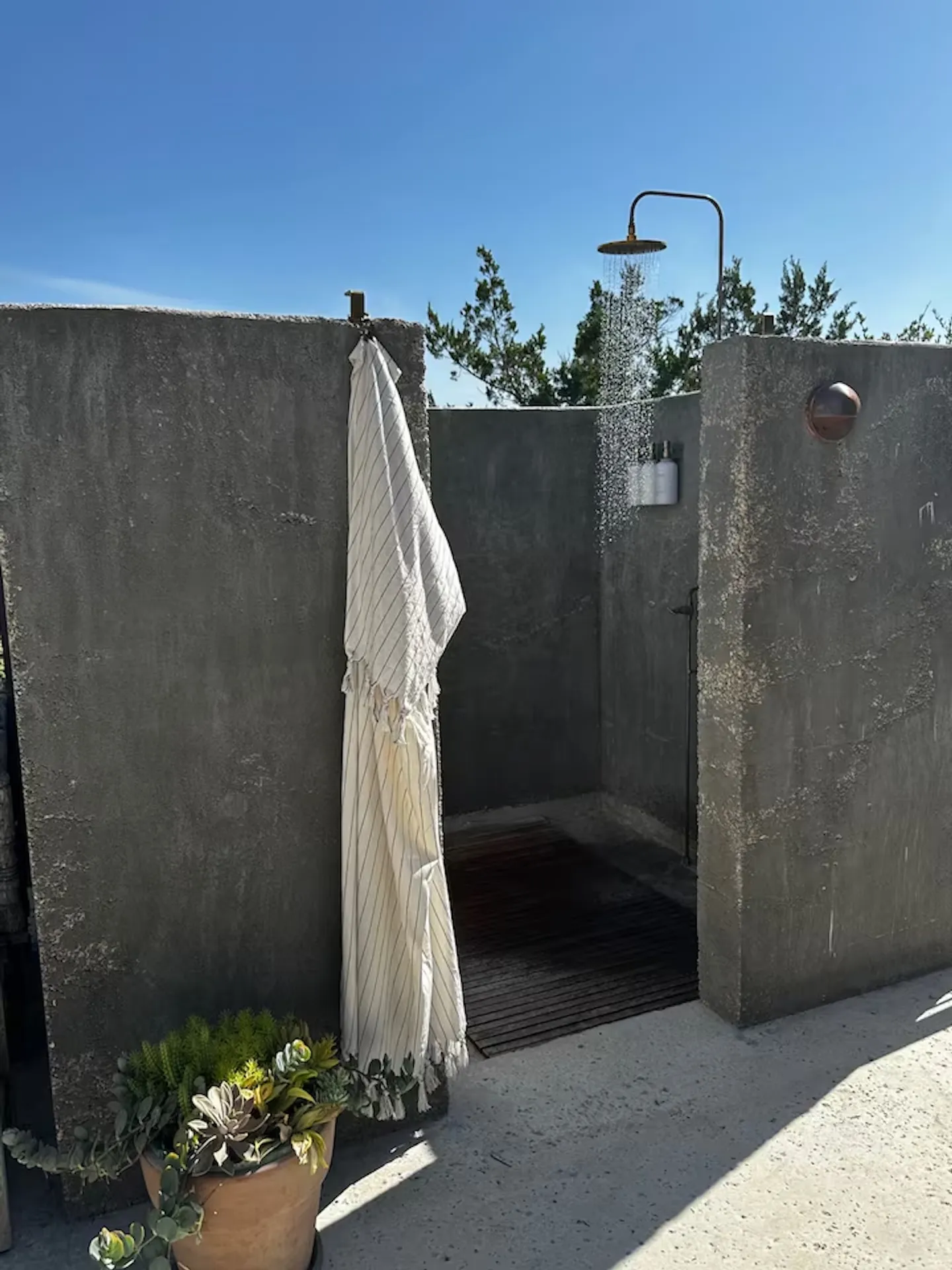 Outdoor shower enclosure made of textured concrete walls with running water from an overhead showerhead under a clear blue sky.