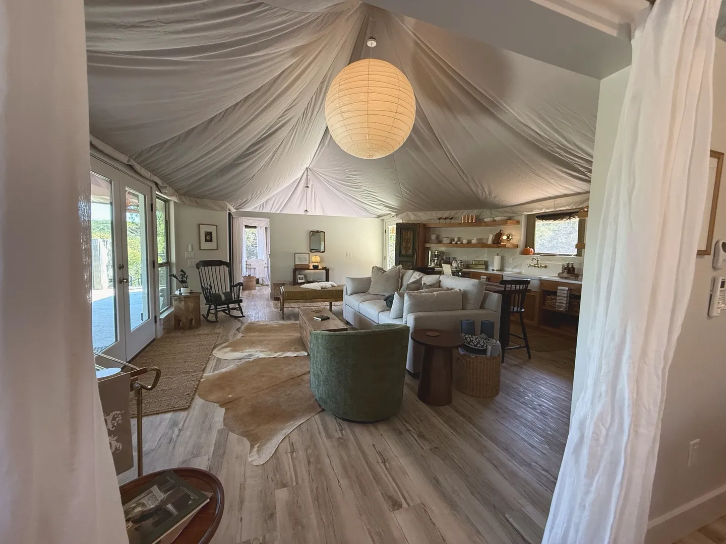 Open living area with a fabric-draped ceiling, large spherical paper lantern, sofa, armchair, and open kitchen in the background.