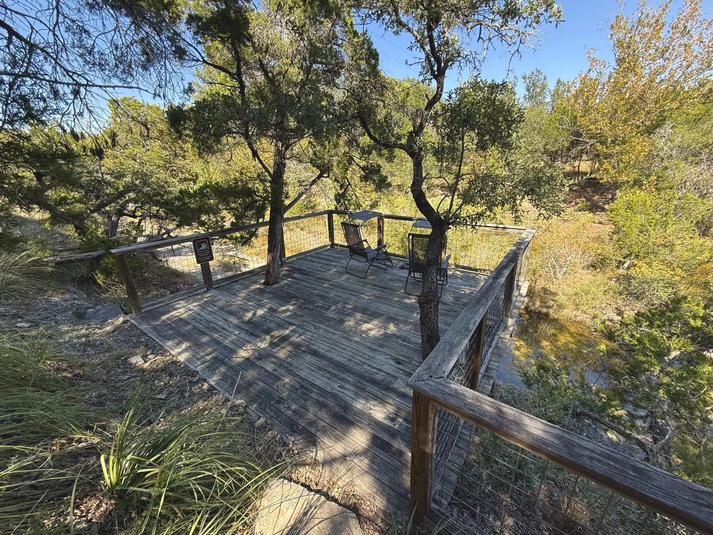 Elevated wooden deck nestled among trees with two lounge chairs and railings overlooking a wooded creek area.
