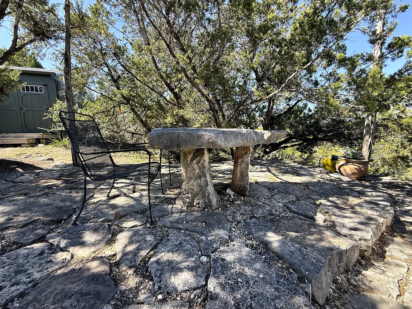 A rustic stone table and black metal mesh chairs on a flagstone patio shaded by trees.