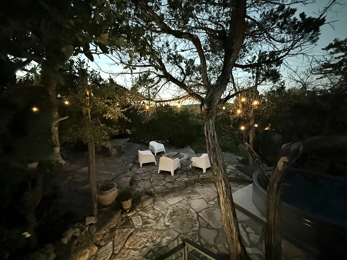Outdoor stone patio at twilight with a fire pit, modern white chairs, string lights strung between trees, and a stock tank pool or hot tub to the side.