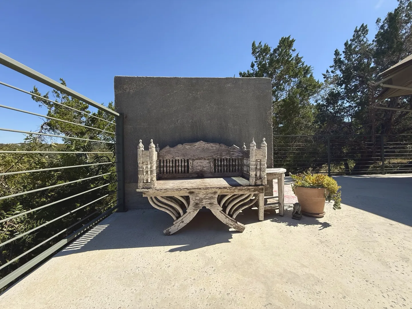 Ornate wooden bench with carved posts on an outdoor deck against a gray concrete wall under a blue sky.