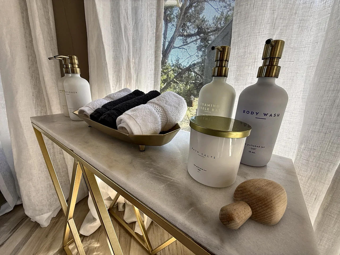 Close-up of a marble-topped console table with bath amenities including rolled towels, bottled soaps, and bath salts in front of sheer curtains.