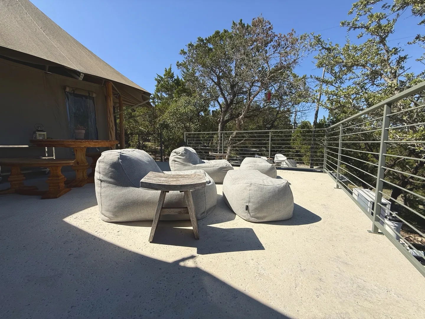 Outdoor deck area outside a safari-style tent with cushioned lounge beanbag seating, a wooden side table, and metal railing overlooking trees.