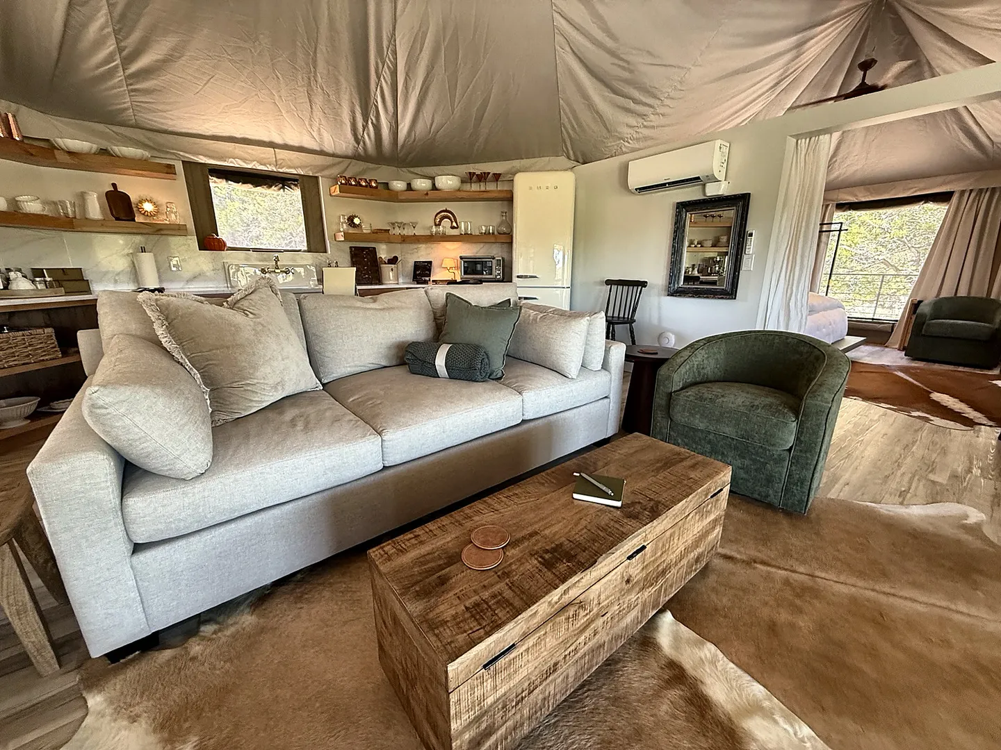 Living area inside a safari-style tent with a fabric ceiling, featuring a grey sofa, wooden coffee table, cowhide rug, and open kitchen shelves behind it.
