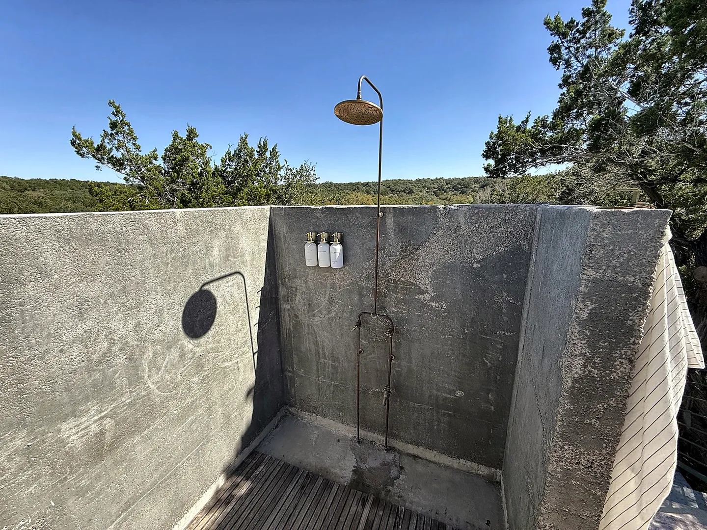 Outdoor shower enclosed by concrete-style half-walls with a slatted wooden floor and distant trees under a clear blue sky.