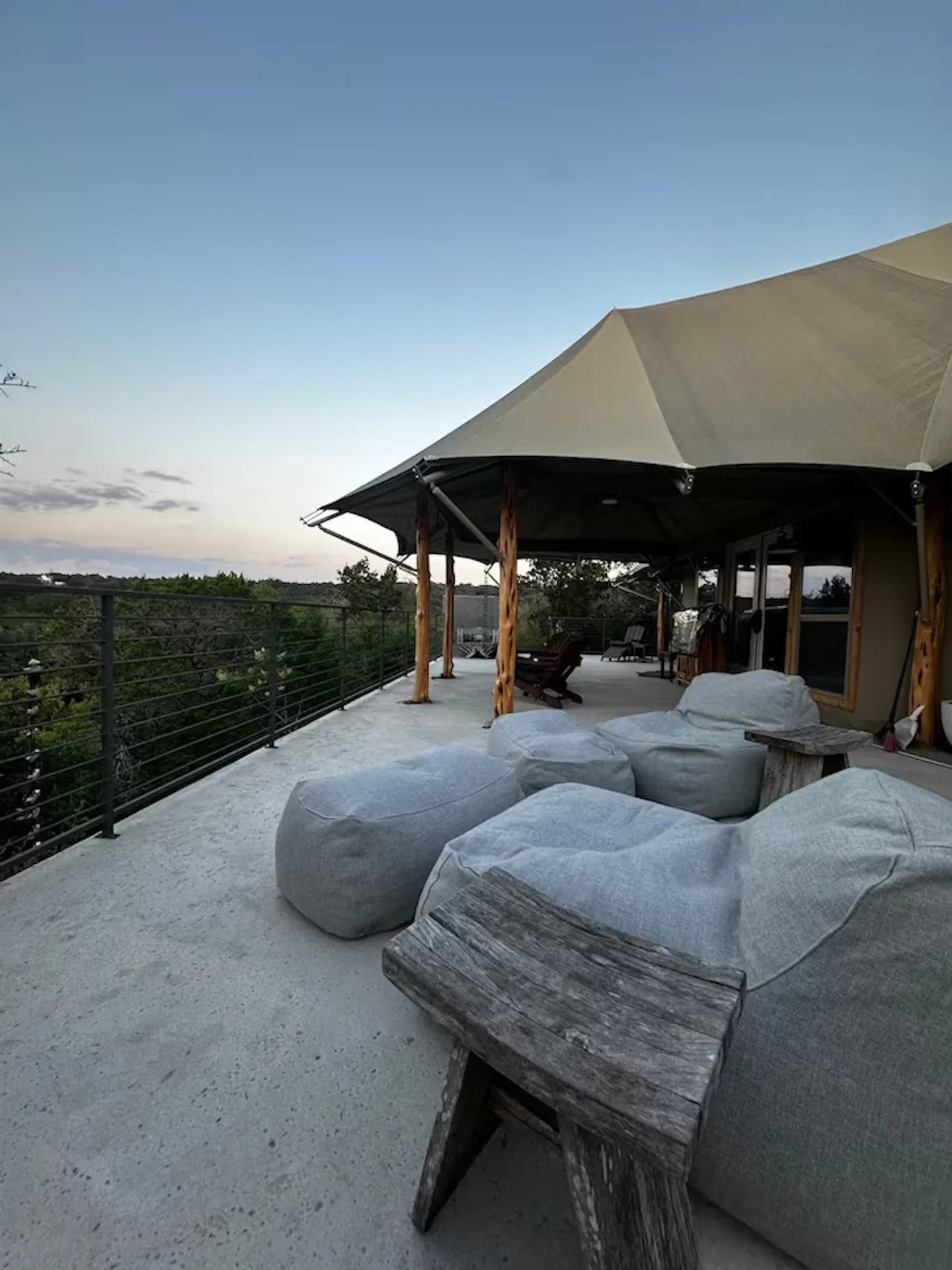 Outdoor concrete deck beside a canvas safari-style tent structure with bean bag loungers, rustic wood side tables, and a metal railing overlooking trees at twilight.
