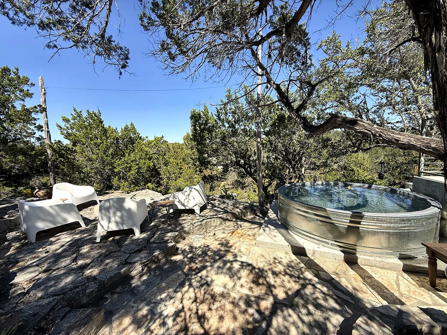 Outdoor stone patio with white lounge chairs and a round galvanized stock tank pool surrounded by trees under a clear blue sky.