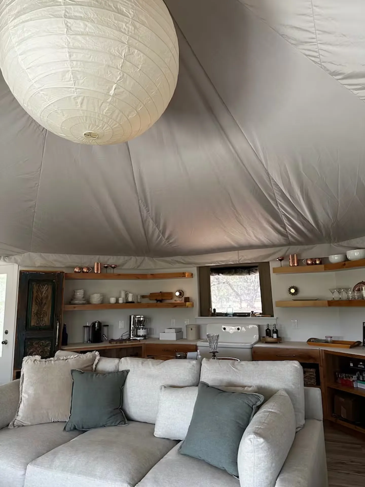 Interior of a yurt featuring a living area with a sectional sofa and an open kitchenette with wooden shelving beneath a conical fabric ceiling.