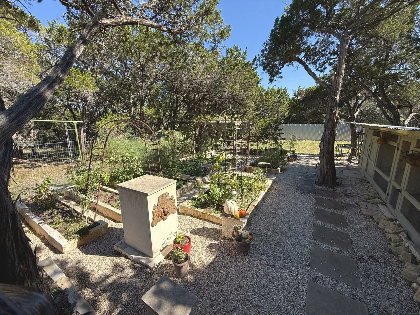 Outdoor garden area with raised plant beds, gravel pathways, stepping stones, and an enclosed coop structure.