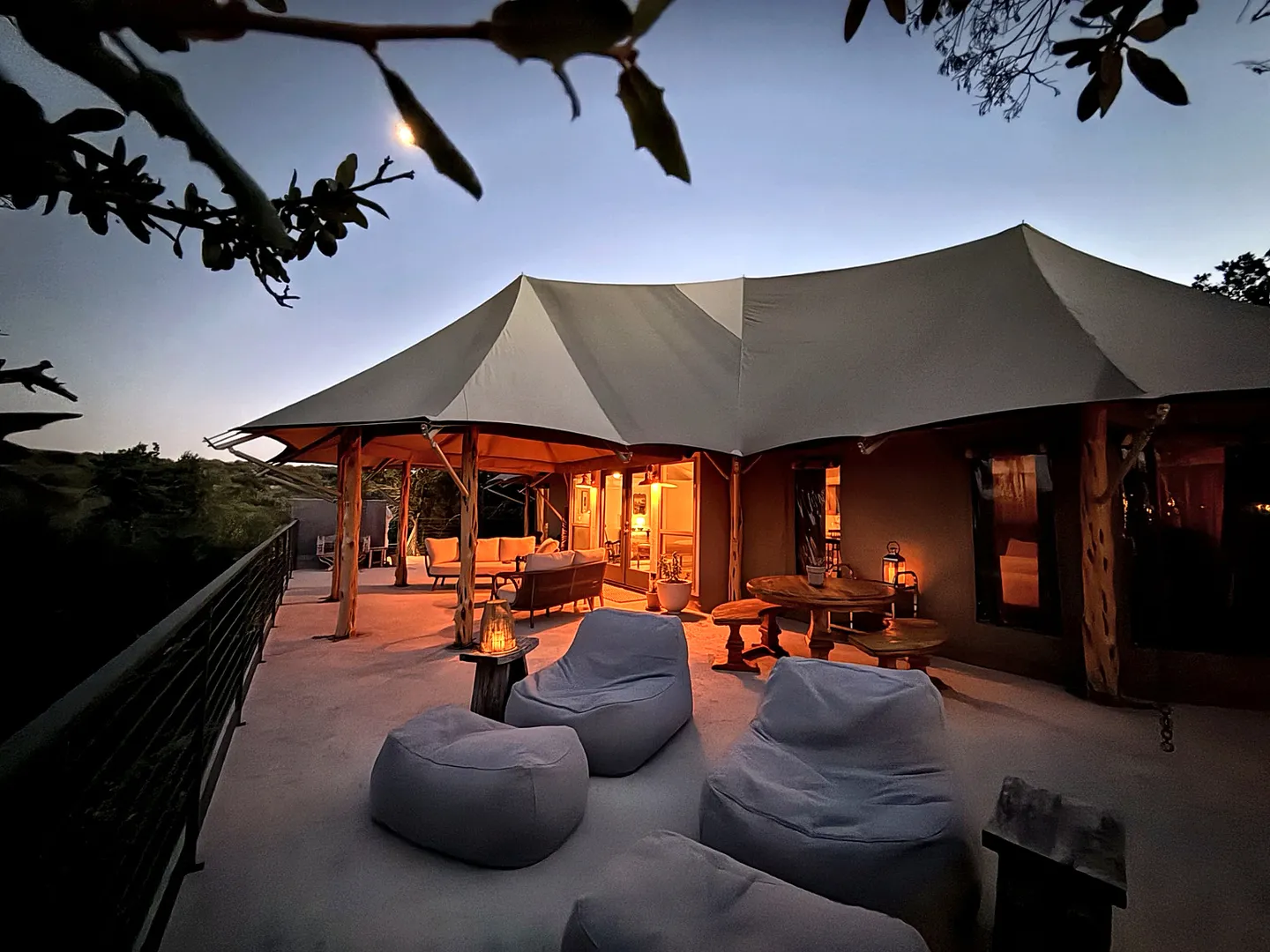 Outdoor deck at dusk with bean bag loungers, a wooden dining table, and seating under the canopy of a safari-style tent structure.