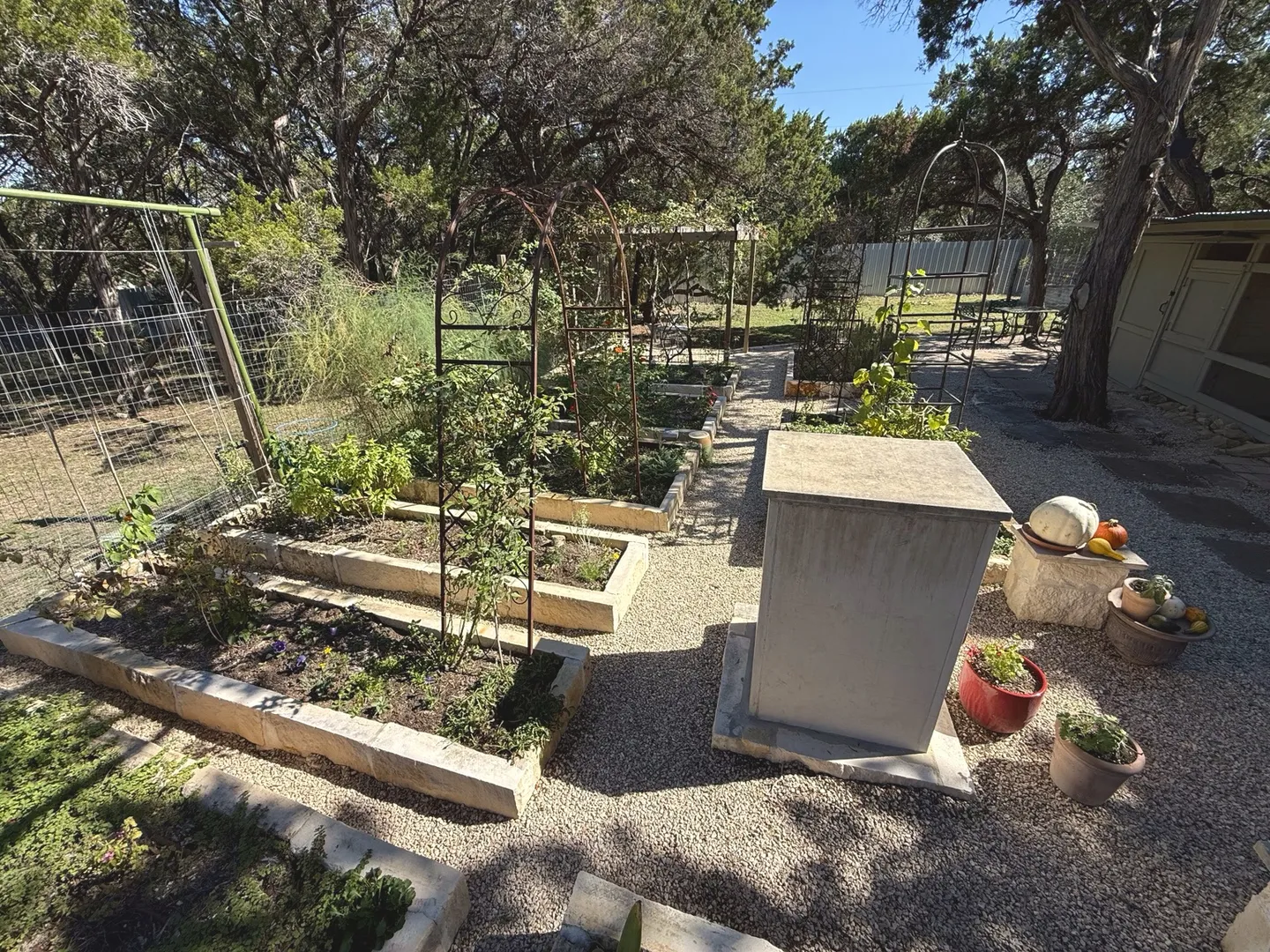 Outdoor garden area featuring stone raised garden beds, gravel pathways, metal trellises, and surrounding trees under a clear blue sky.
