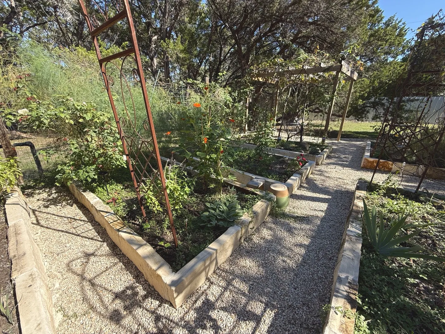 Outdoor garden area featuring raised stone planting beds, metal trellises, gravel paths, and a wooden arbor.