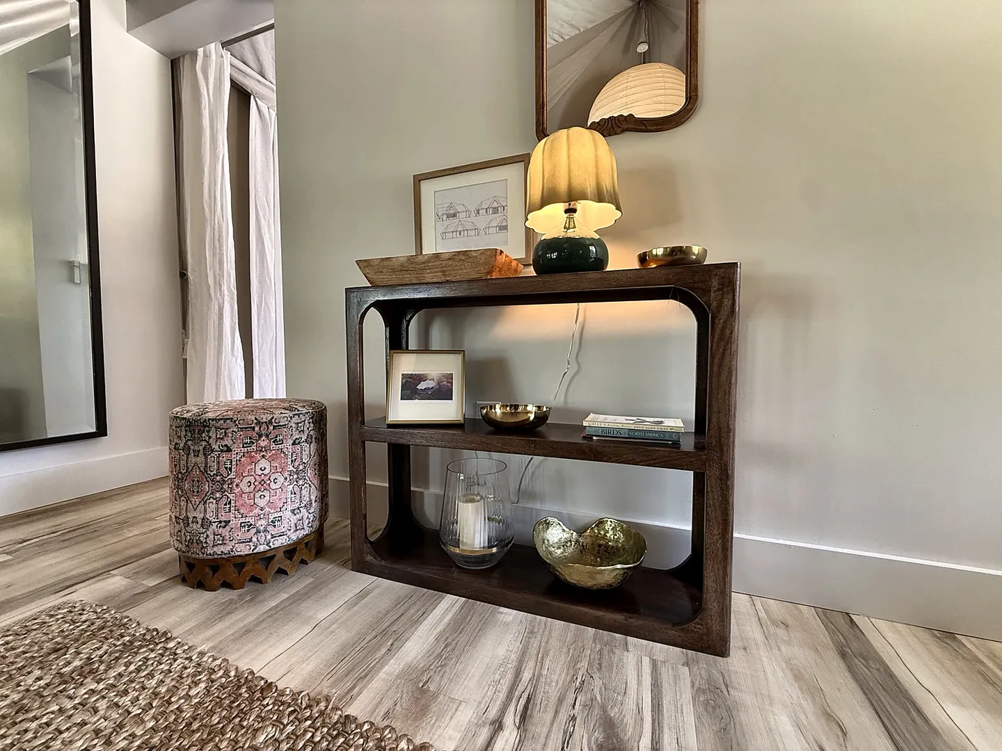 A wooden console table decorated with a lamp, bowls, and framed art against a light wall, next to a patterned ottoman stool.